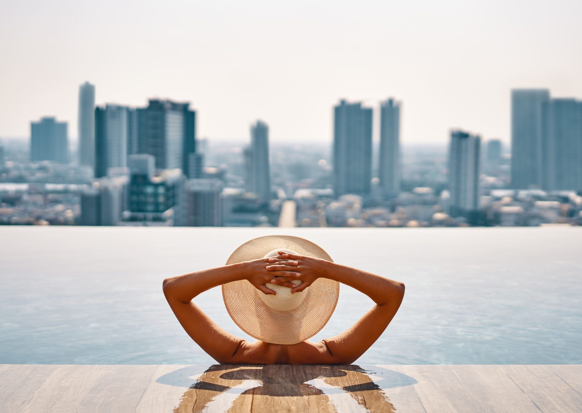 Image of a woman with a hat on, enjoying the cityscape with a view from her own private pool.